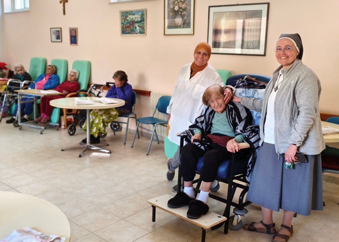 An elderly resident of Our Lady of Sorrows smiling while seated in a communal garden with a Sister and a lay volunteer