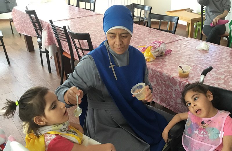 A caregiver at Hogar Niño Dios smiling and assisting a young resident in a wheelchair during an outdoor activity