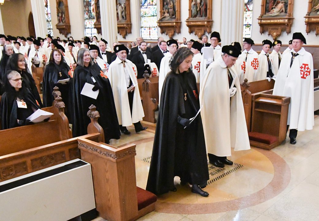 Knights and Dames of the Order processing into a cathedral during an investiture ceremony, wearing white capes bearing the Jerusalem Cross
