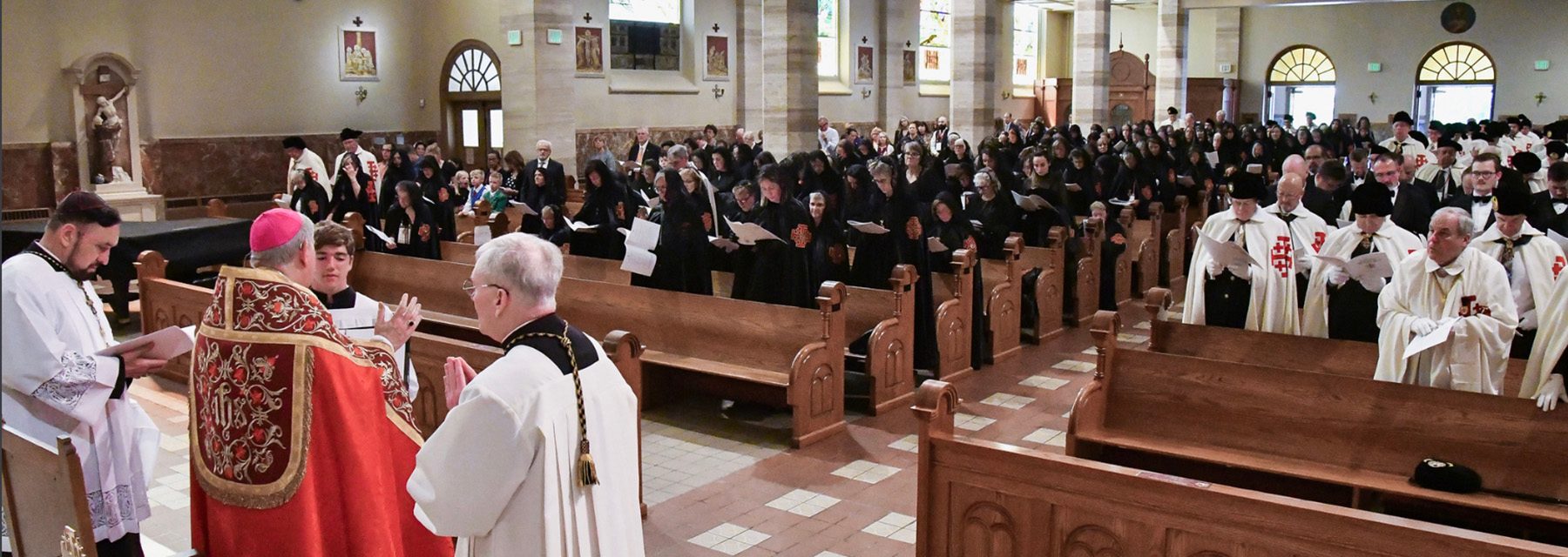 New Knights and Dames kneeling at the altar during an investiture ceremony, receiving their capes bearing the Jerusalem Cross, with clergy and existing members looking on